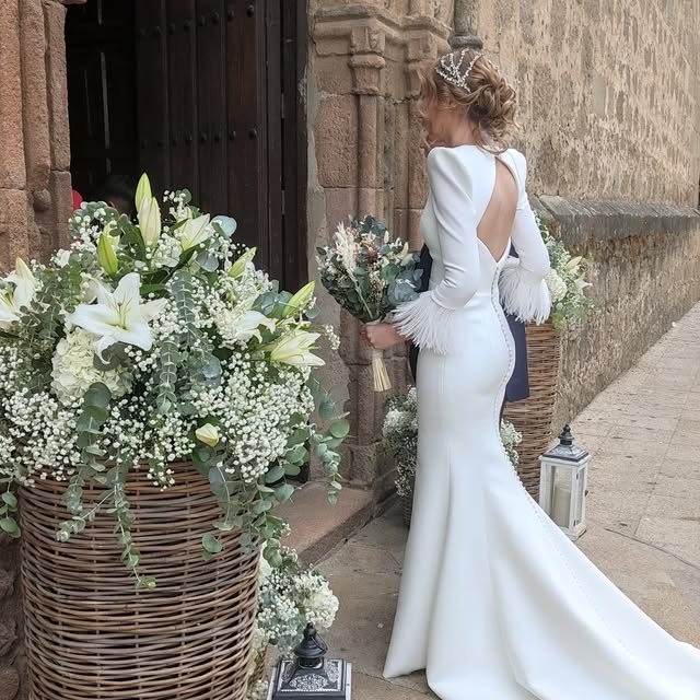 Novia de espaldas junto a una gran cesta de lirios blancos y paniculata, a la puerta de una iglesia